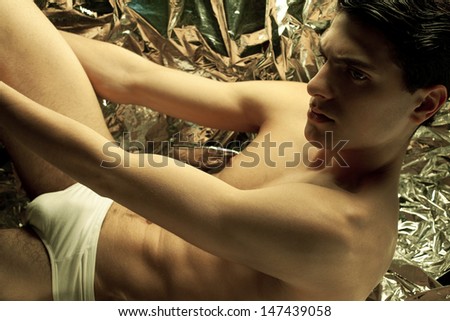 Beautiful (handsome) muscular male model with nice abs and posing over wrinkled silver foil background. Perfect skin and body. White underwear. Vogue style. Fashion studio portrait