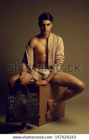 Male high fashion concept. Portrait of a handsome male model sitting on a wooden cube in trendy cardigan and underwear, holding leather bag. Perfect skin & haircut. Vogue style. Studio shot