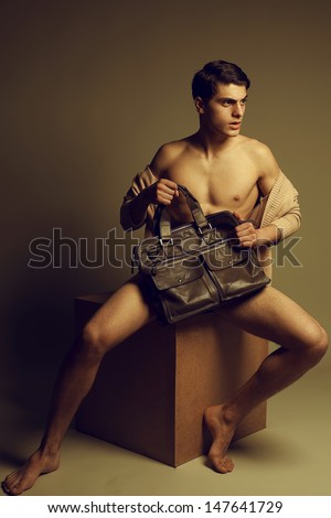 Male high fashion concept. Portrait of a handsome male model sitting on a wooden cube in trendy cardigan, holding leather bag. Perfect skin & haircut. Vogue style. Copy-space. Studio shot