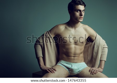 Male high fashion concept. Portrait of a handsome male model sitting on a wooden cube in trendy cardigan and stylish underwear. Perfect skin & haircut. Vogue style. Copy-space. Studio shot