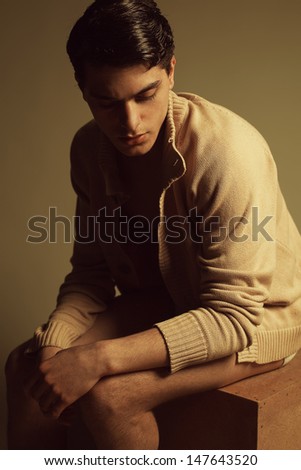 Male high fashion concept. Portrait of a handsome male model sitting on a wooden cube in trendy cardigan and looking down. Perfect skin & haircut. Vogue style. Studio shot