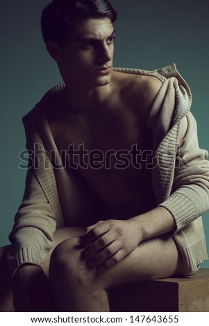 Male high fashion concept. Portrait of a handsome male model sitting on a wooden cube in trendy cardigan and looking somewhere. Perfect skin & haircut. Vogue style. Studio shot