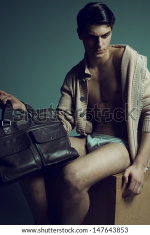 Male high fashion concept. Portrait of a handsome male model sitting on a wooden cube in trendy cardigan & underwear, holding leather bag. Perfect skin & haircut. Vogue style. Copy-space. Studio shot