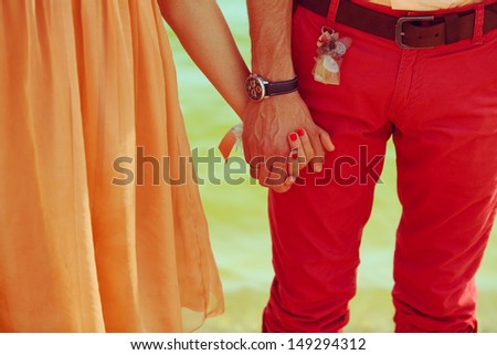 Close Up Holding Hands Together Of Young Couple Of Hipsters In Love. Stylish Trendy Clothes. Peach And Red Colors. Vintage Style. Outdoor Shot.