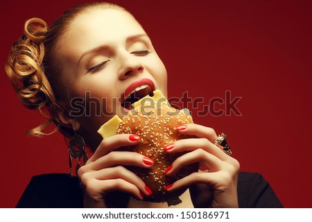 Unhealthy eating. Junk food concept. Guilty pleasure. Portrait of happy fashionable model holding burger & eating over red background. Perfect hair, skin, make-up & manicure. Copy-space. Studio shot