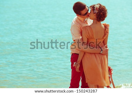 Happy honeymoon (vacation) concept. Young happy married hipsters in trendy clothes standing and kissing over blue ocean (sea) background. Sunny summer day. Copy-space. Outdoor shot