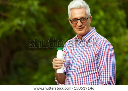 Healthy food concept. Portrait of a mature (old) man in trendy casual shirt and glasses posing in the park and holding a bottle with milk. Sunny summer day. Copy-space. Outdoor shot