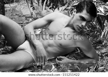 Beautiful (handsome) muscular male model with nice abs in sitting and posing over wrinkled silver foil background. Perfect skin and body. White underwear. Vogue style. Black & white studio portrait