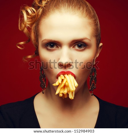 Unhealthy eating. Junk food concept. Arty portrait of fashionable young woman holding (eating) fried potato in her mouth and posing over red background. Close up. Studio shot