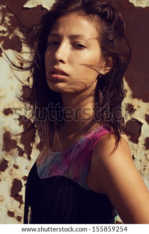 Portrait of a young beautiful model wearing trendy black dress and posing over rust stains background. Sunny & windy weather. Vogue style. Close up. Outdoor shot