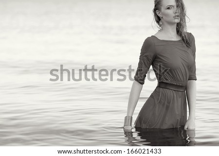 Fashion portrait of a young model with wet long ginger curly hair in wet grey dress posing in the water. Perfect skin and make-up. Copy-space. Black and white (monochrome) outdoor shot