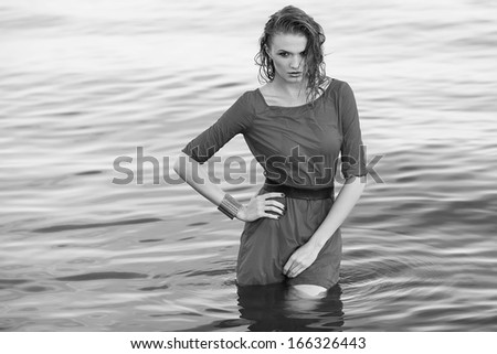 Fashion portrait of a young model with wet long ginger curly hair in wet grey dress posing in the water. Perfect skin and make-up. Black and white (monochrome) outdoor shot