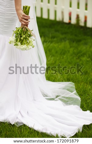Bride\'s hands with wedding bouquet of beige flowers over white wooden fence and green lawn. Trendy dress with vapory veil. Country (vintage) style. Copy-space. Outdoor shot