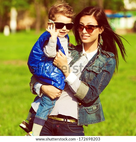 Hi & bye concept. Portrait of fashionable baby boy and his gorgeous mother in trendy sunglasses walking in the street. Mom hugging son. Sunny spring day. Outdoor shot