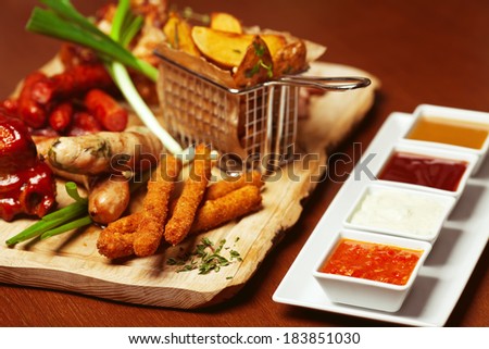 Fried cheese sticks with small meat sausages served with scallion and dried herbs on wooden cutting board with a set of marinara, mayonnaise, honey-mustard sauces and ketchup. Copy-space. Indoor shot