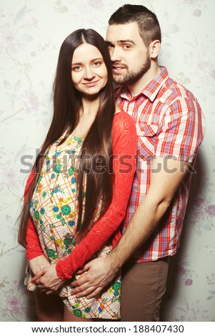 Stylish pregnancy concept: portrait of couple of hipsters (husband and wife) in trendy clothes (shirt, dress, cardigan and jeans) hugging & expecting their child. Studio shot