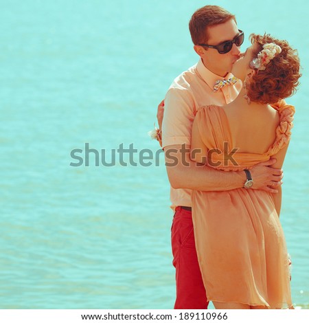 Stylish honeymoon concept. Young happy married couple of hipsters in trendy clothes, eyewear and accessories standing and kissing on the beach. Sunny summer day. Copy-space. Outdoor shot