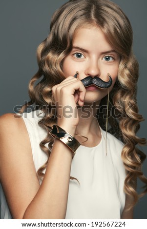 Teen star concept. Portrait of beautiful teen girl with long blond glossy hair posing in trendy clothing with luxurious golden accessories. Girl hiding behind mustache. Hipster style. Studio shot