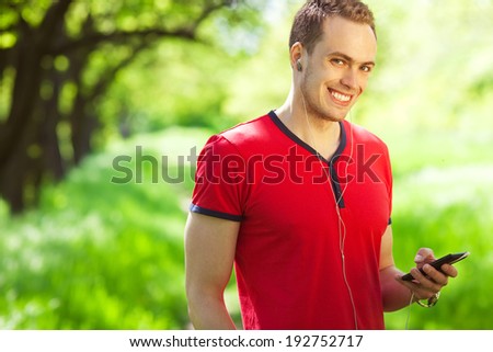 Gadget freak concept. Portrait of a young muscular man in red t-shirt and jeans walking with cellphone in the park. Great white shiny smile. Sunny weather. Close up. Copy-space. Outdoor shot
