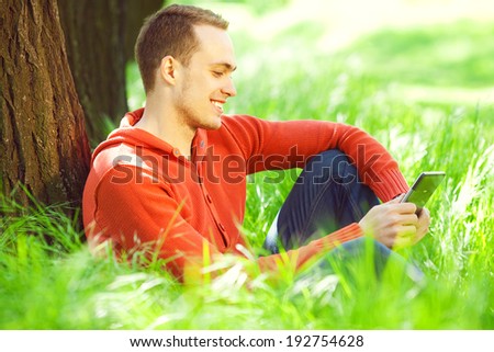 Gadget freak concept. Portrait of smiling young man in casual clothing reading e-book, sitting in green grass under tree in the park. Great white shiny smile. Close up. Sunny weather. Outdoor shot