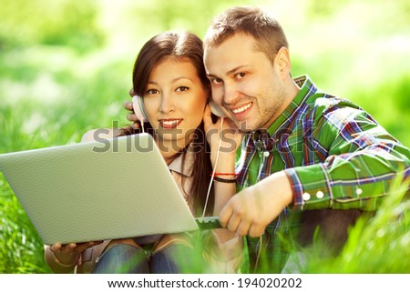 Happy couple of hipsters in trendy casual clothing using a laptop (with earphones) in park (forest, garden) and looking sweet and in love. Perfect shiny white smiles. Outdoor shot
