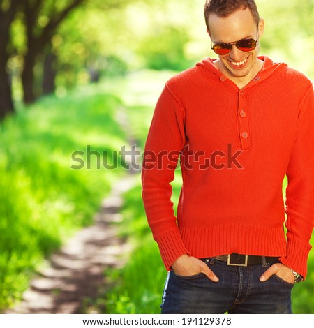 Eyewear concept. Portrait of a  shy young muscular man in trendy clothing and sunglasses laughing and staying near road in the park (forest). Sunny spring (autumn) weather. Copy-space. Outdoor shot