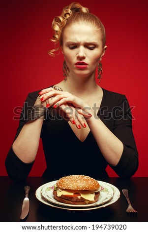 Junk food concept. Portrait of unhappy luxurious red-haired model in black cocktail dress trying to eat burger. Perfect hair, skin, make-up and manicure. Golden accessories. Studio shot