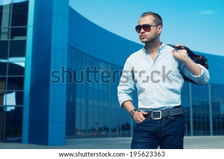 Portrait of attractive man in trendy casual clothing with leather bag and sunglasses posing over shopping mall. Sunny spring weather with blue sky. Outdoor shot