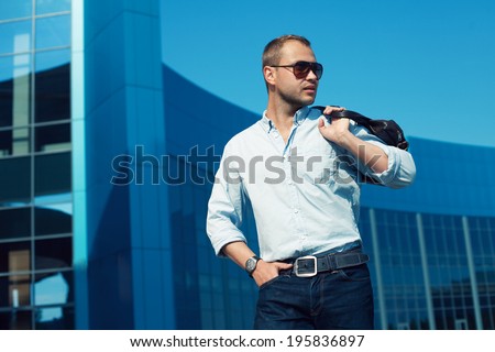 Men Shopping concept. Portrait of attractive smiling man in trendy casual clothing with leather bag and sunglasses posing over shopping mall. Sunny spring weather with blue sky. Outdoor shot