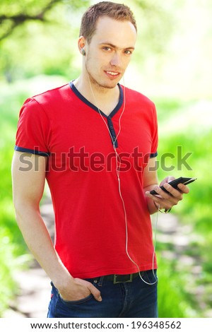 Portrait of a smiling young man in red t-shirt using smartphone in the park. Handsome muscular guy in casual clothing. Sunny summer day. Outdoor shot