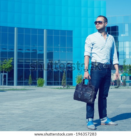 Metrosexual Man Concept. Portrait of attractive man in trendy casual clothing with leather bag and sunglasses posing over shopping mall. Sunny spring weather with blue sky. Outdoor shot