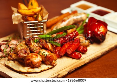 German food concept. Set of fried meat and small meat sausages served with scallion and dried herbs on wooden cutting board with fried potato pieces and set of different sauces. Close up