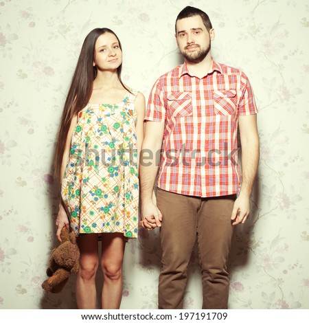 Stylish pregnancy concept: portrait of couple of hipsters (husband and wife) in trendy clothes (shirt, dress, cardigan and jeans) holding brown teddy bear & smiling. Vintage (retro) style. Studio shot