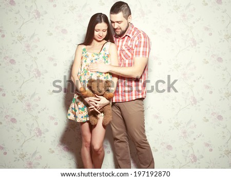 Stylish pregnancy concept: portrait of couple of hipsters (husband and wife) in trendy clothes (shirt, dress, jeans) holding teddy bear and smiling. Vintage (retro) style. Copy-space. Studio shot