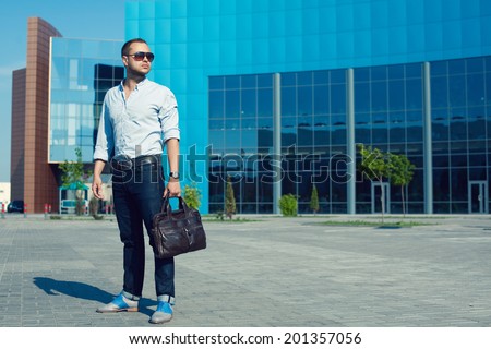 Hipster Man Concept. Portrait of attractive guy in trendy casual clothing with leather bag and sunglasses posing over shopping mall. Sunny summer weather with blue sky. Outdoor shot