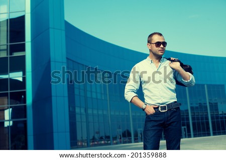 Men Shopping concept. Profile portrait of attractive smiling man in trendy casual clothing with sunglasses and leather bag posing over shopping mall. Sunny spring weather with blue sky. Outdoor shot