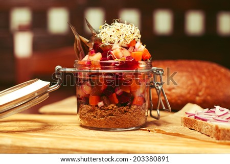 Mason jar with pieces of vegetables (carrot, onion, capers, red beet), anchovy, brown bread crumbles and seedlings of lucerne served with parchment paper on wooden board in restaurant. Close up