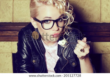 Eyewear concept. Portrait of a funny hipster blonde girl with great hairdo in trendy glasses and black leather jacket eating cake in vintage restaurant. Duck face emotion. Close up. Indoor shot