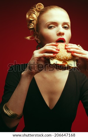 Fashion & Gluttony Concept. Portrait of luxurious red-haired model in black cocktail dress eating burger over red background. Perfect hair, skin, make-up and manicure. Golden accessories. Studio shot