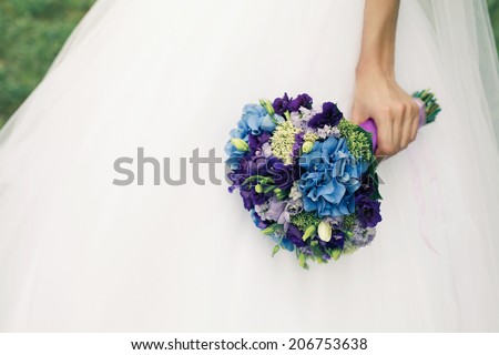 Bride\'s hand with wedding bouquet of blue flowers over white vapory dress and green lawn. Vintage style. Close up. Copy-space. Outdoor shot