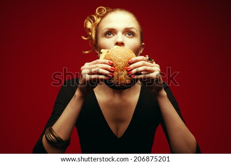 Fashion & Gluttony Concept. Portrait of luxurious red-haired model in black cocktail dress eating burger over red background. Funny curls, perfect skin, manicure. Golden accessories. Studio shot