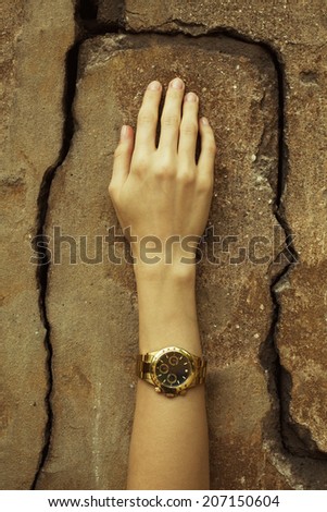 Luxurious accessory concept. Young woman\'s hand wearing a golden watch with golden strap over cracked stone wall. Perfect manicure. Vintage style. Close up. Outdoor shot