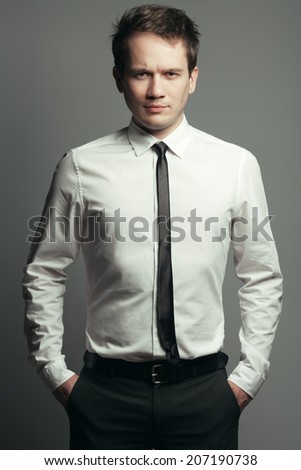Portrait of serious handsome young man in casual white shirt, black tie, leather belt, gray pants posing over grey background. Hands in trousers pockets. Stylish haircut. Hipster style. Studio shot