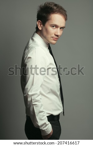 Portrait of serious handsome young man in casual white shirt, black tie, leather belt, gray pants posing over grey background. Hands in trousers pockets. Stylish haircut. Hipster style. Studio shot