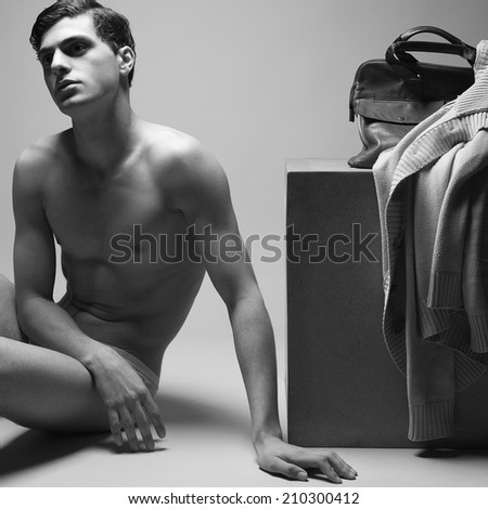 Male high fashion concept. Portrait of handsome male model sitting near wooden cube in trendy underwear. Cardigan & leather bag on cube. Perfect skin & haircut. Vogue style. Black & white studio shot