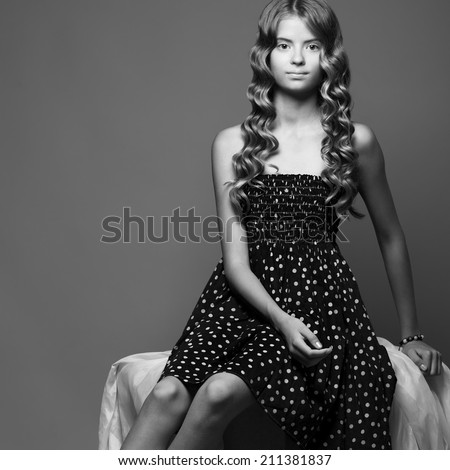 Modern Mona Lisa concept. Beautiful girl in black dress sitting on white cloth over gray background. Healthy skin and glossy long curly hair. Copy-space. Black and white (monochrome) studio shot