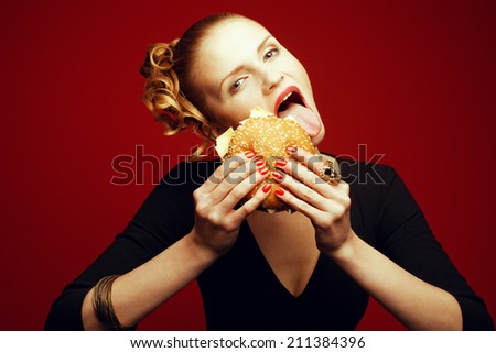 Fashion & Gluttony Concept. Happy red-haired model in black cocktail dress sticking out tongue & eating burger over red background. Perfect skin, make-up & manicure. Golden accessories. Studio shot