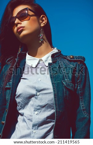 Eyewear concept. Fashion portrait of stylish brunette in trendy sunglasses and jeans jacket. Luxurious vintage earrings. Urban grunge style. Outdoor shot