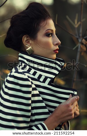 Emotive profile portrait of Hollywood diva, film star walking in the park. Evening time. Sunny spring weather. Young woman in luxurious trendy striped jacket. Golden earrings. Close up. Outdoor shot