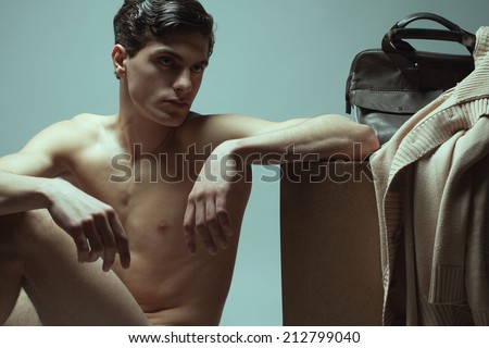 Male high fashion concept. Portrait of a handsome young man sitting near wooden cube. Cardigan and leather bag on cube. Perfect skin & haircut. Wet hair. Vogue style. Studio shot